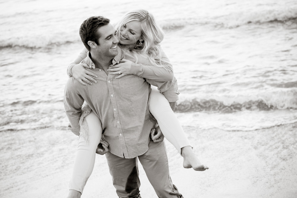 A man gives a woman a piggyback ride on a beach, both smiling and looking at each other, with waves in the background. Jessica Hill Photography - Wedding Photographer - Oregon