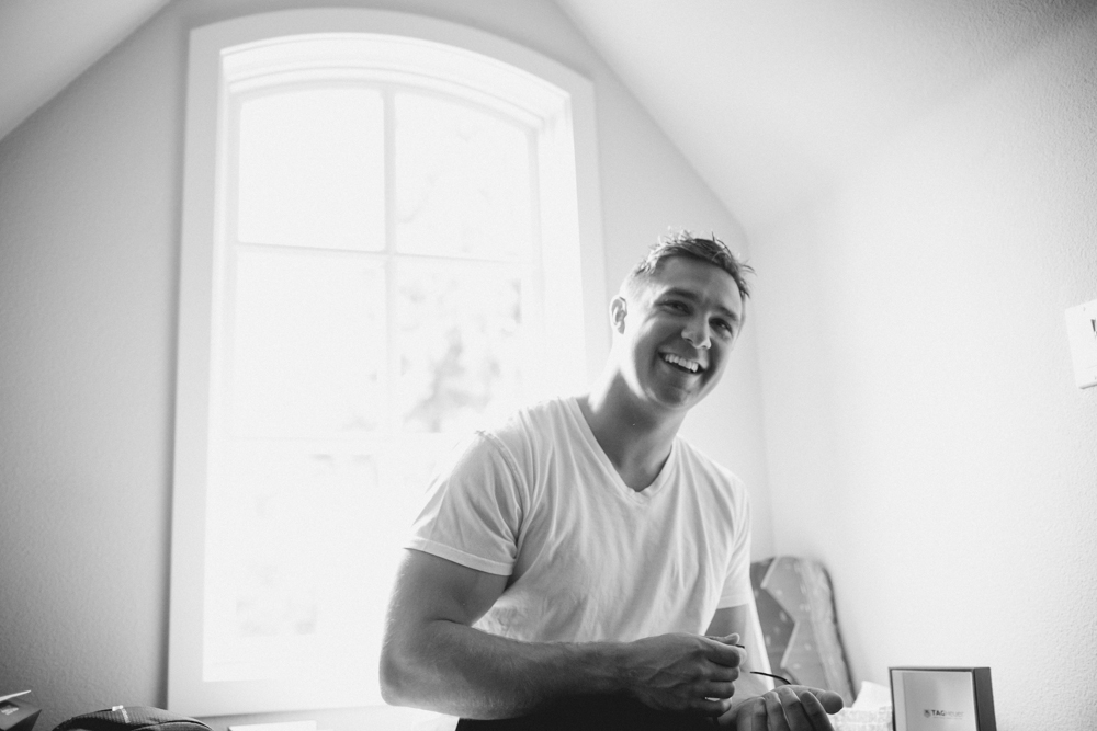 A man in a white t-shirt smiles and looks toward the camera while standing in a bright room with a large arched window behind him. The image is in black and white. Jessica Hill Photography - Wedding Photographer - Oregon