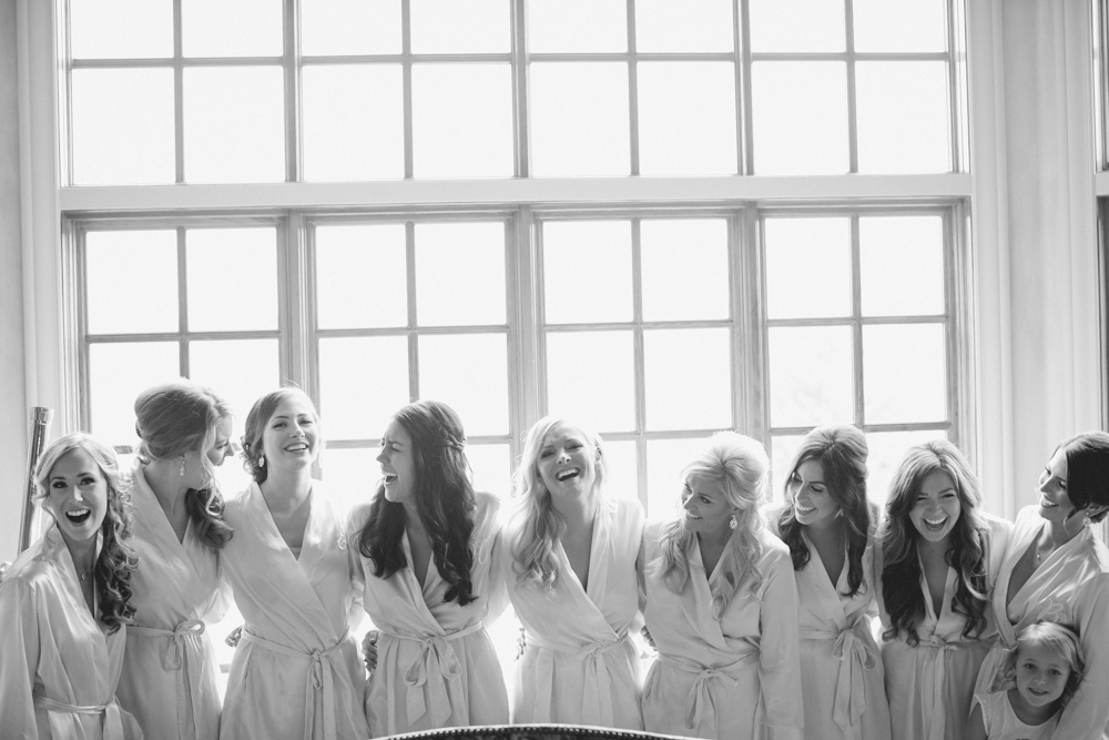 A group of women and one child, all wearing matching robes, stand together in front of large windows, smiling and laughing. The image is bright and joyful, suggesting a celebratory or pre-wedding moment. Jessica Hill Photography - Wedding Photographer - Oregon