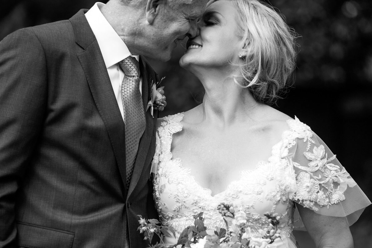 A bride and groom smile and touch noses lovingly on their wedding day. The bride wears a lace wedding dress and holds a bouquet, while the groom wears a suit and tie. The photo is in black and white. Jessica Hill Photography - Wedding Photographer - Oregon