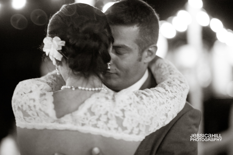 A bride and groom embrace closely while dancing, with the bride's back to the camera. String lights are blurred in the background. The image is in black and white. Jessica Hill Photography - Wedding Photographer - Oregon
