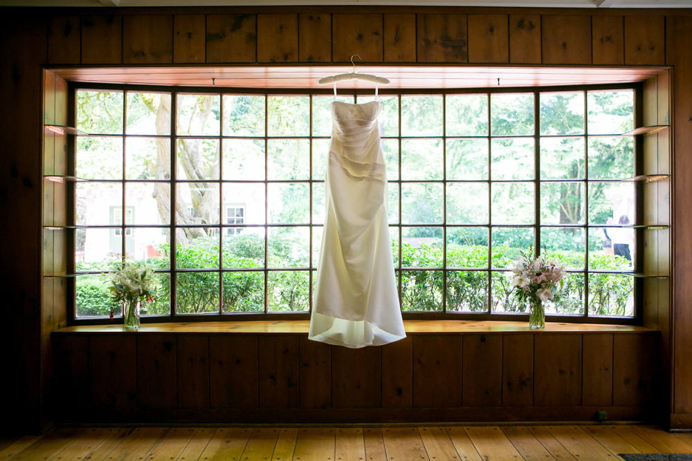 A white strapless wedding dress hangs on a hanger in front of a large window with wooden trim, flanked by two vases of flowers on a wooden bench. Jessica Hill Photography - Wedding Photographer - Oregon