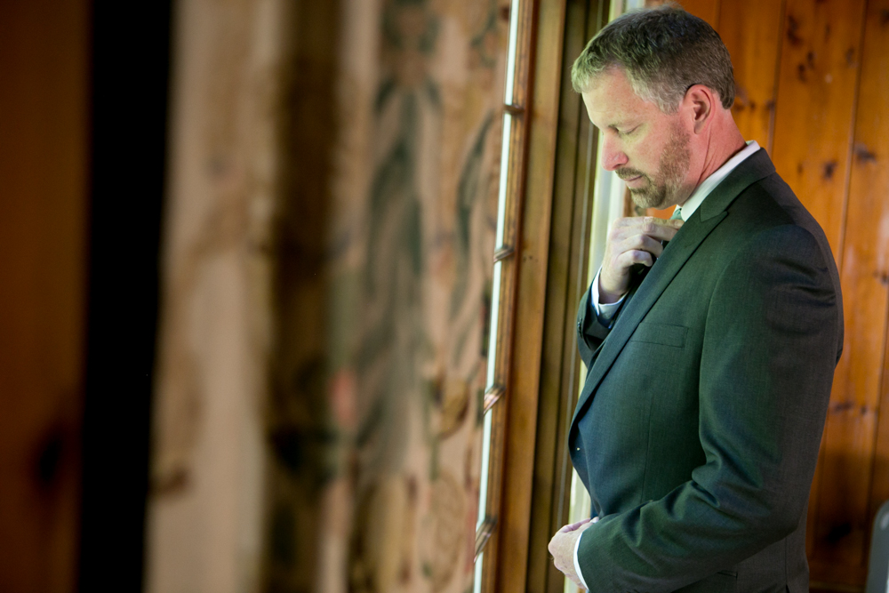 A man in a suit adjusts his tie while standing near a window with wooden walls and patterned curtains in the background. Jessica Hill Photography - Wedding Photographer - Oregon