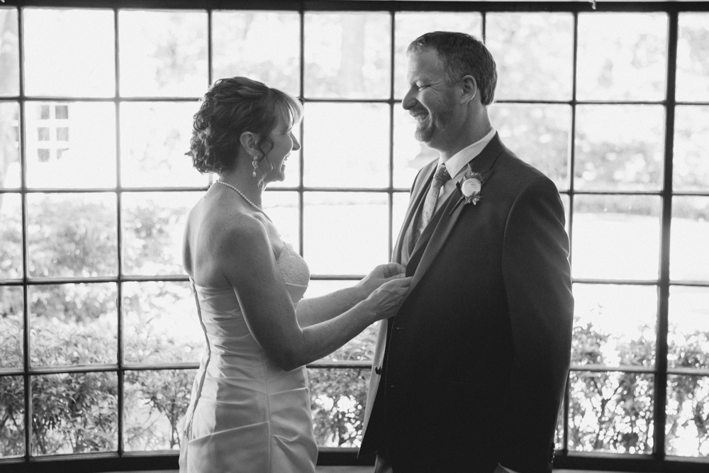 A bride and groom stand facing each other, smiling, in front of a large window with a garden view. Jessica Hill Photography - Wedding Photographer - Oregon
