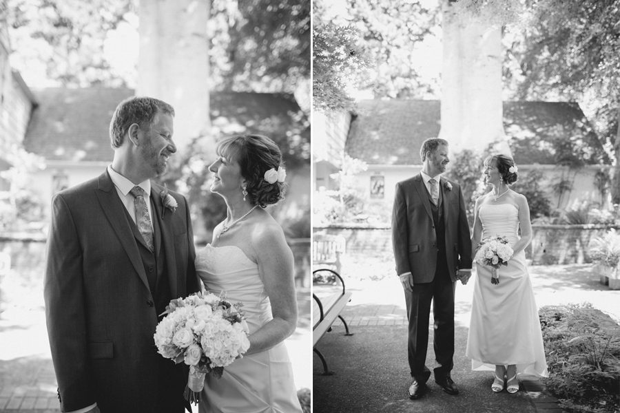 A bride and groom stand outdoors in formal attire, facing each other and smiling, with trees and a building in the background. The image is in black and white. Jessica Hill Photography - Wedding Photographer - Oregon