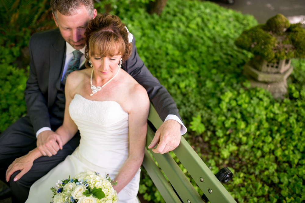 A bride in a white dress and a groom in a suit sit closely together on a green bench, surrounded by green foliage. Jessica Hill Photography - Wedding Photographer - Oregon
