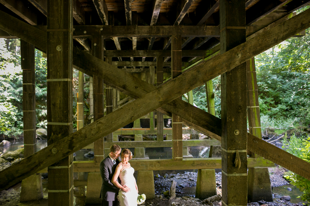 A couple stands together on a stone path surrounded by tall, dense trees and lush greenery in a forest. Jessica Hill Photography - Wedding Photographer - Oregon