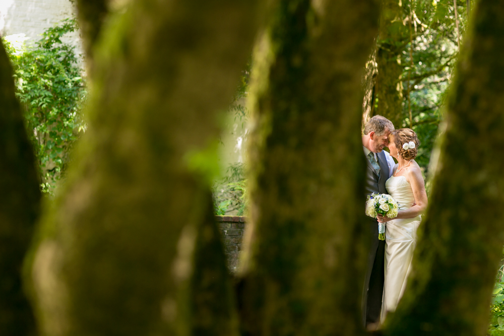 A bride and groom stand closely together outdoors, partially obscured by tree trunks, with greenery in the background. Jessica Hill Photography - Wedding Photographer - Oregon