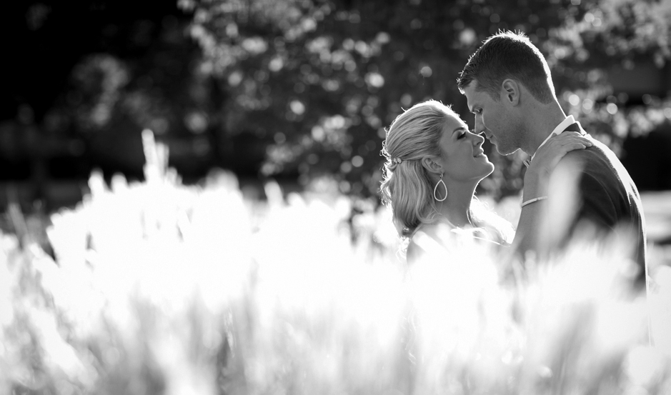 A couple stands close together outdoors, facing each other with soft expressions, surrounded by blurred, sunlit foliage. Jessica Hill Photography - Wedding Photographer - Oregon