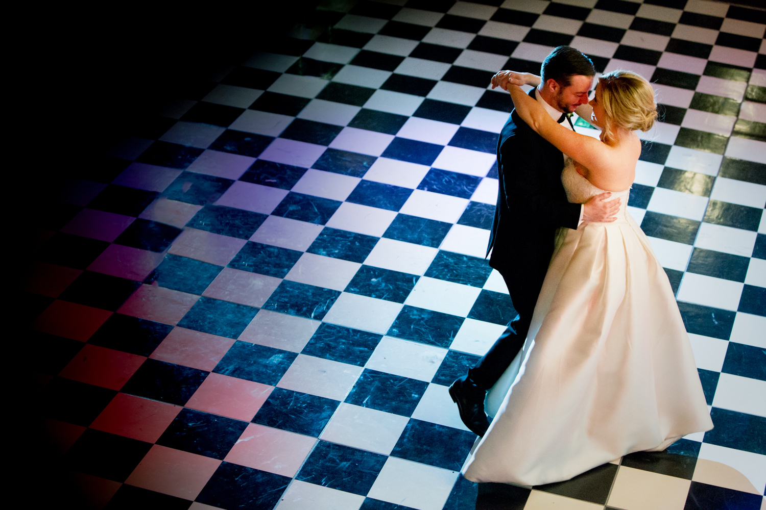 A bride and groom share a first dance on a black-and-white checkered floor, embracing each other closely under soft lighting. Jessica Hill Photography - Wedding Photographer - Oregon
