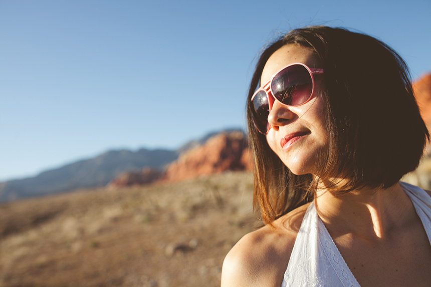 A woman with short brown hair and pink sunglasses smiles outdoors in a sunlit, rocky desert landscape with mountains in the background. Jessica Hill Photography - Wedding Photographer - Oregon