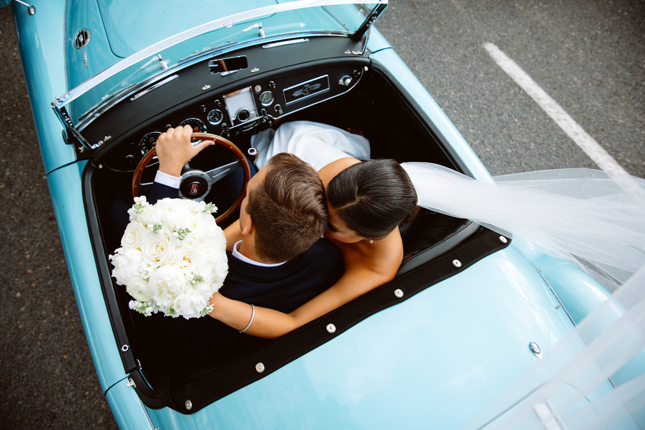 Overhead view of a bride and groom sitting in a vintage blue convertible, with the bride holding a bouquet of white flowers. Jessica Hill Photography