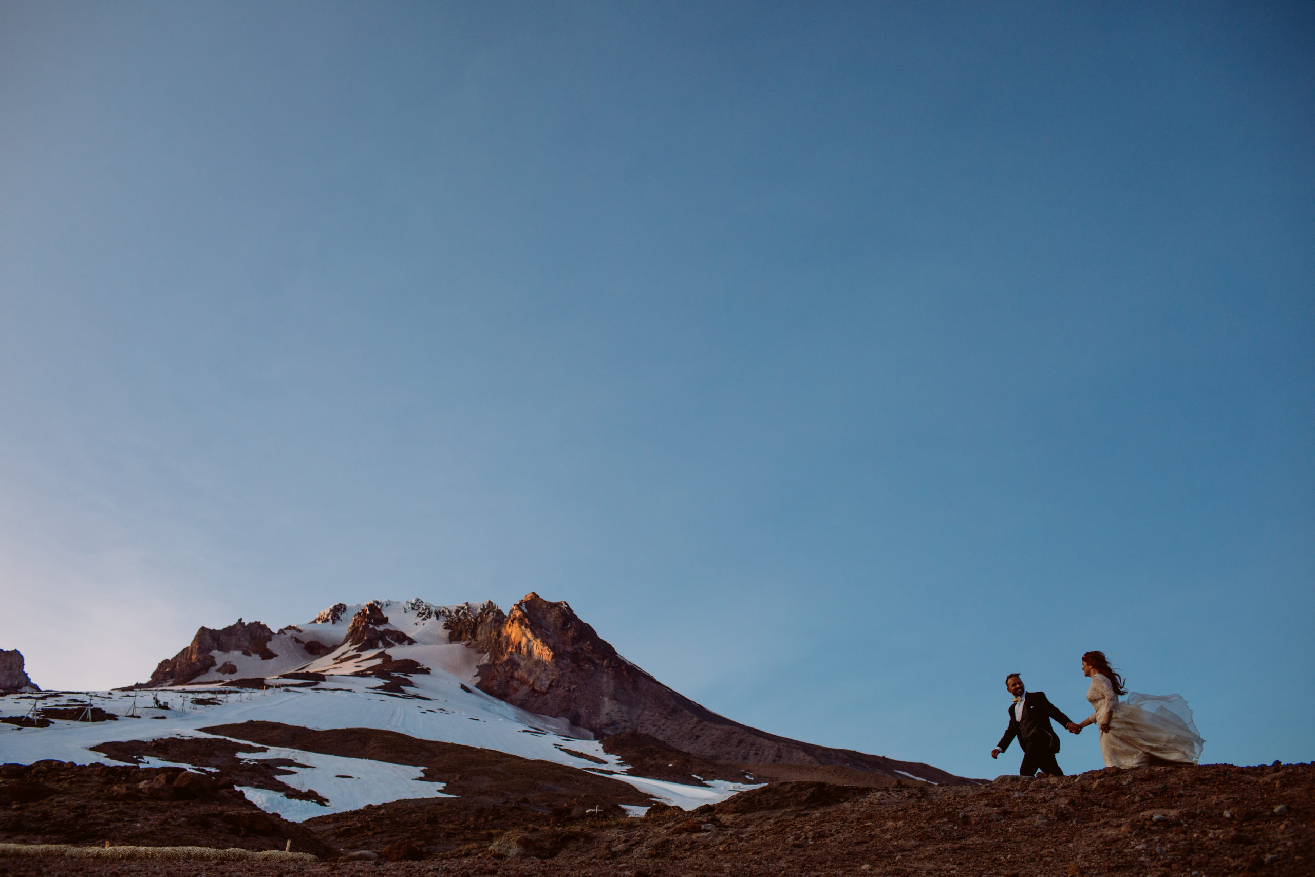 A couple in wedding attire holds hands and walks along a rocky slope with a snow-capped mountain in the background under a clear sky. Jessica Hill Photography