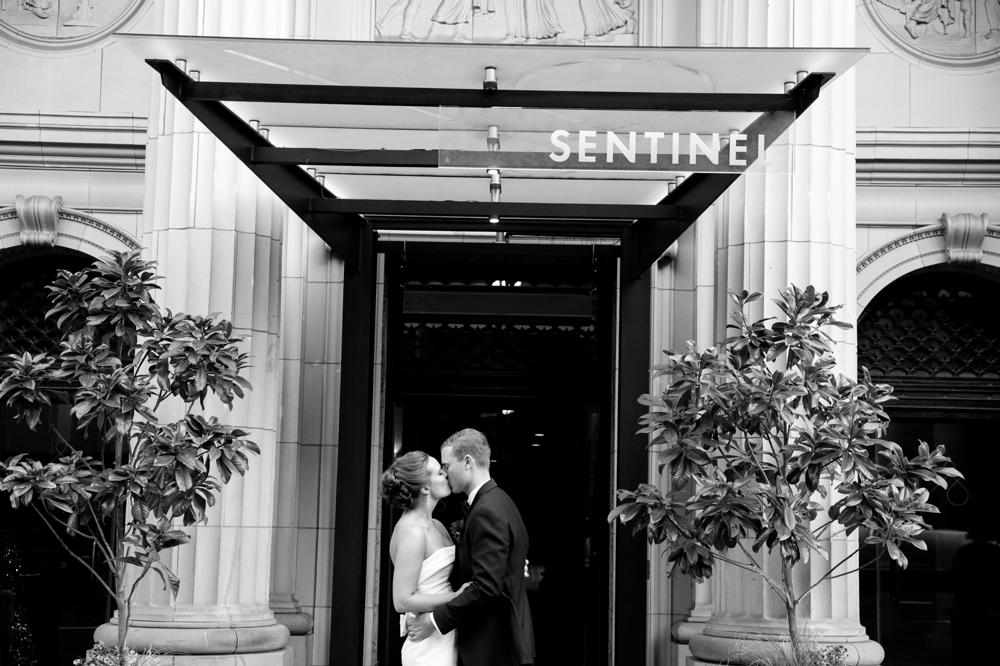 A bride and groom stand close together outside the entrance of a building labeled "Sentinel," framed by columns and plants. Jessica Hill Photography
