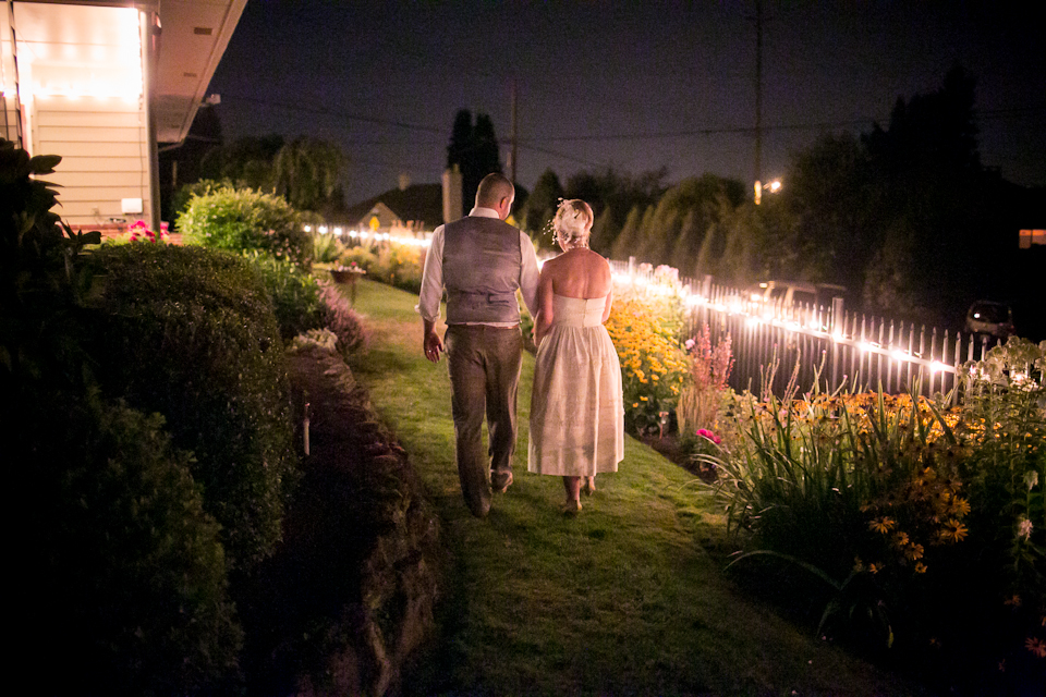 A couple in formal attire walks hand in hand along a garden path at night, illuminated by string lights. Jessica Hill Photography - Wedding Photographer - Oregon
