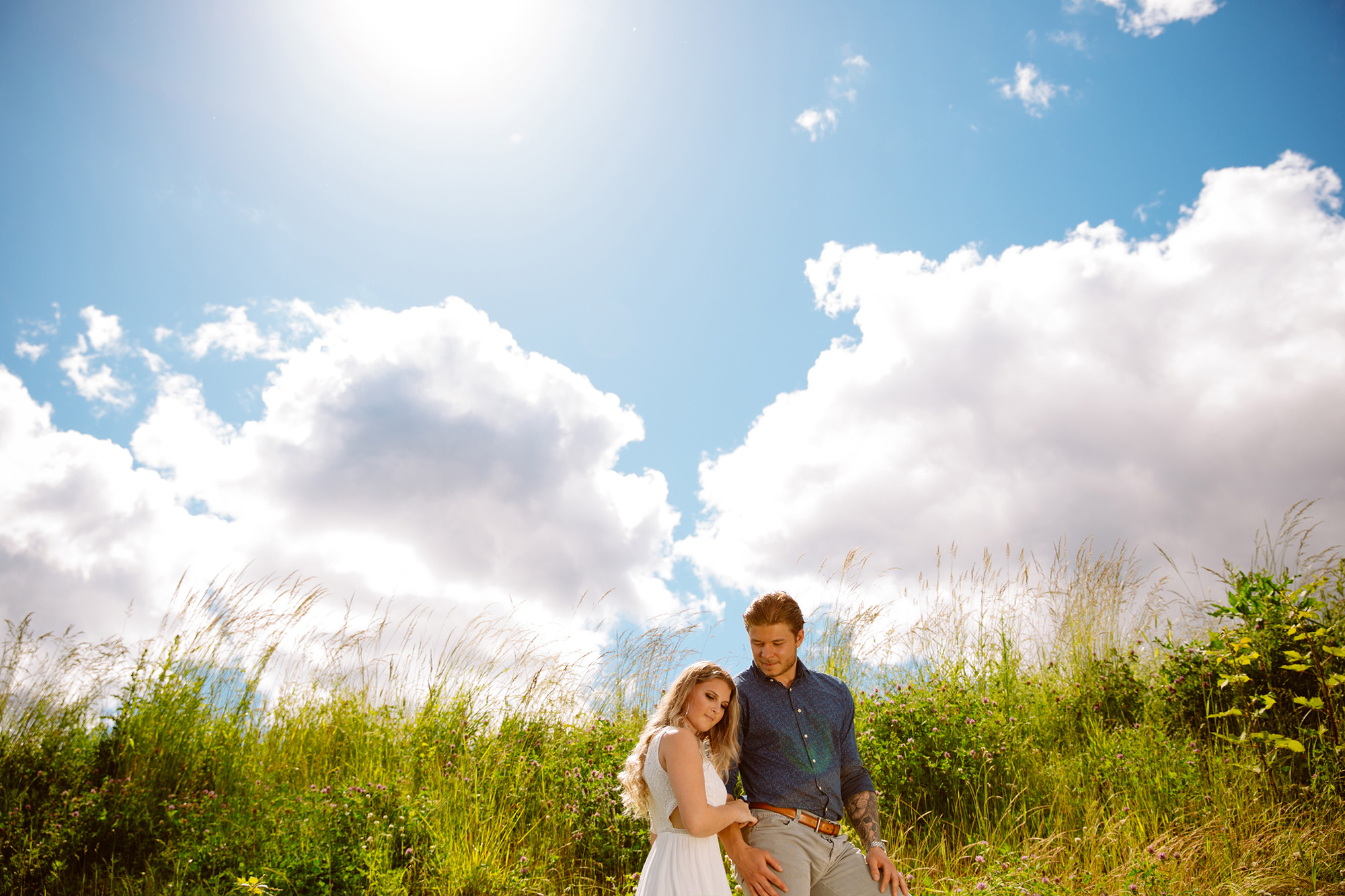 A couple stands closely together in a grassy field under a bright blue sky with fluffy clouds. The woman leans gently on the man’s shoulder, and sunlight shines overhead, creating a warm, serene atmosphere. Jessica Hill Photography - Wedding Photographer - Oregon
