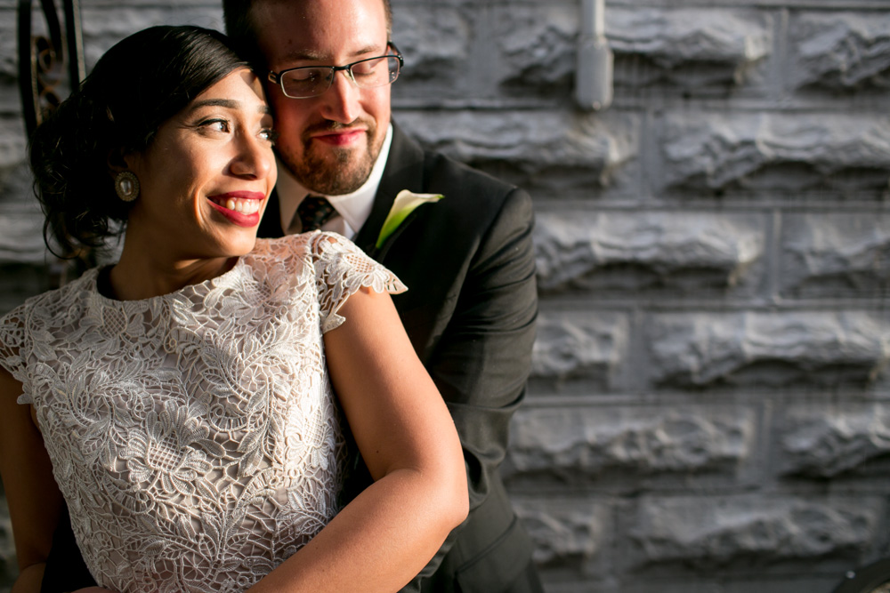 A couple dressed formally embraces and smiles in front of a textured gray brick wall, with sunlight illuminating their faces. Jessica Hill Photography - Wedding Photographer - Oregon