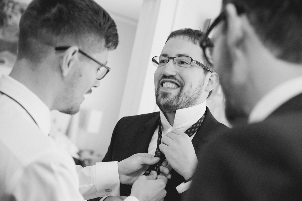 Three men in suits, one helping another adjust his tie, all smiling in a well-lit indoor setting. Jessica Hill Photography - Wedding Photographer - Oregon