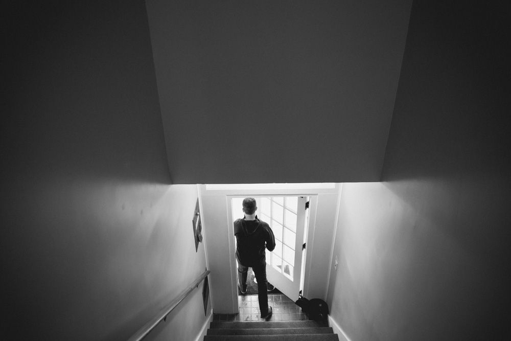 A person seen from behind descends a staircase toward a door with windows, carrying a bag in one hand in a dimly lit hallway. Jessica Hill Photography - Wedding Photographer - Oregon