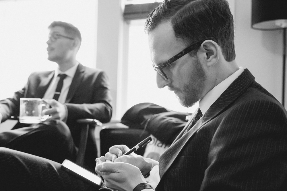 Two men in suits sit indoors; one writes on a notepad while the other holds a mug and looks away. The scene is in black and white. Jessica Hill Photography - Wedding Photographer - Oregon
