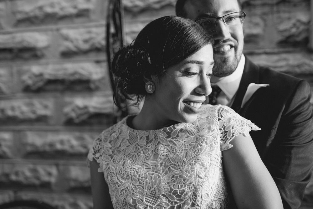 A couple dressed formally, with the woman in a lace dress and the man in a suit, smiling and embracing in front of a brick wall. Jessica Hill Photography - Wedding Photographer - Oregon