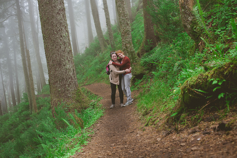 Two people stand hugging and smiling on a dirt trail winding through a lush, green, foggy forest with tall trees and ferns. Jessica Hill Photography - Wedding Photographer - Oregon