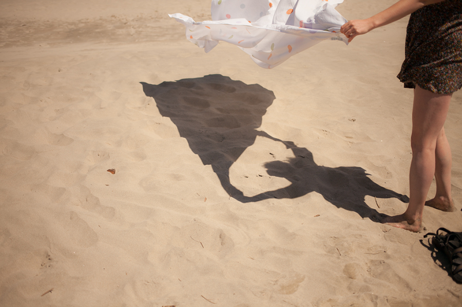 A person stands barefoot on sandy beach, holding up a white patterned sheet that casts a large shadow on the ground. Only their legs and one arm are visible. Jessica Hill Photography - Wedding Photographer - Oregon