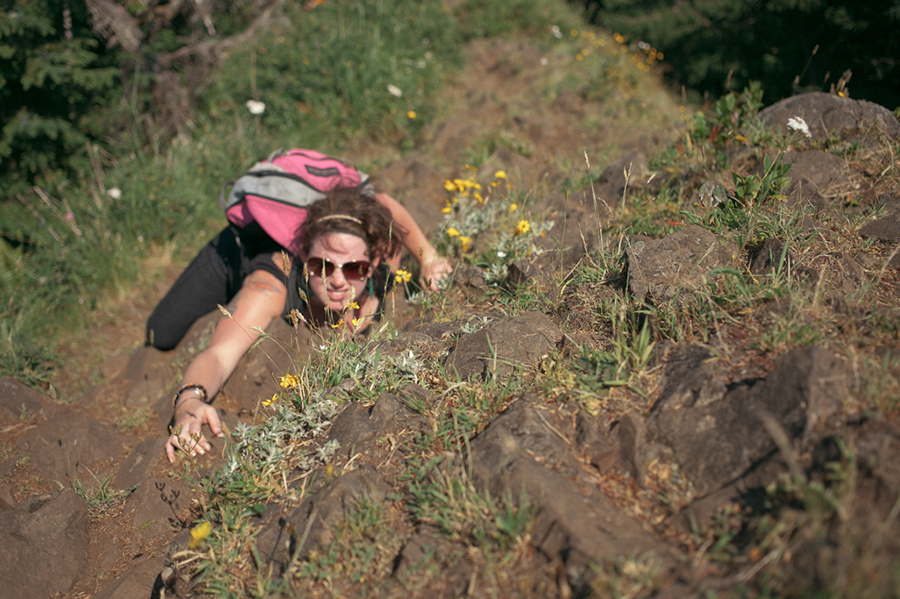 A woman with sunglasses and a pink backpack climbs a steep, rocky hillside covered in grass and yellow flowers, reaching upward with one hand while gripping the rocks with the other. Jessica Hill Photography - Wedding Photographer - Oregon