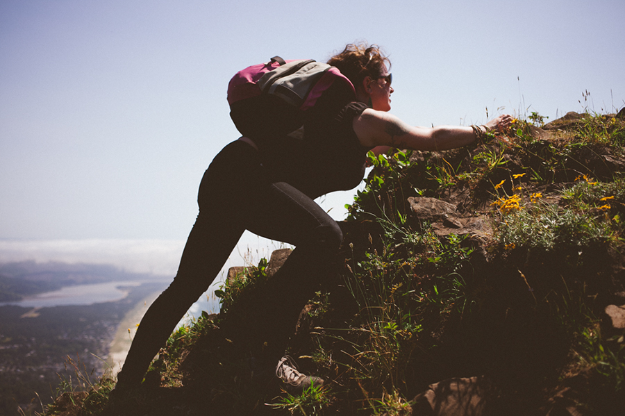 A woman wearing a backpack climbs up a steep, rocky hillside with grass and wildflowers, overlooking a river and landscape below on a sunny day. Jessica Hill Photography - Wedding Photographer - Oregon