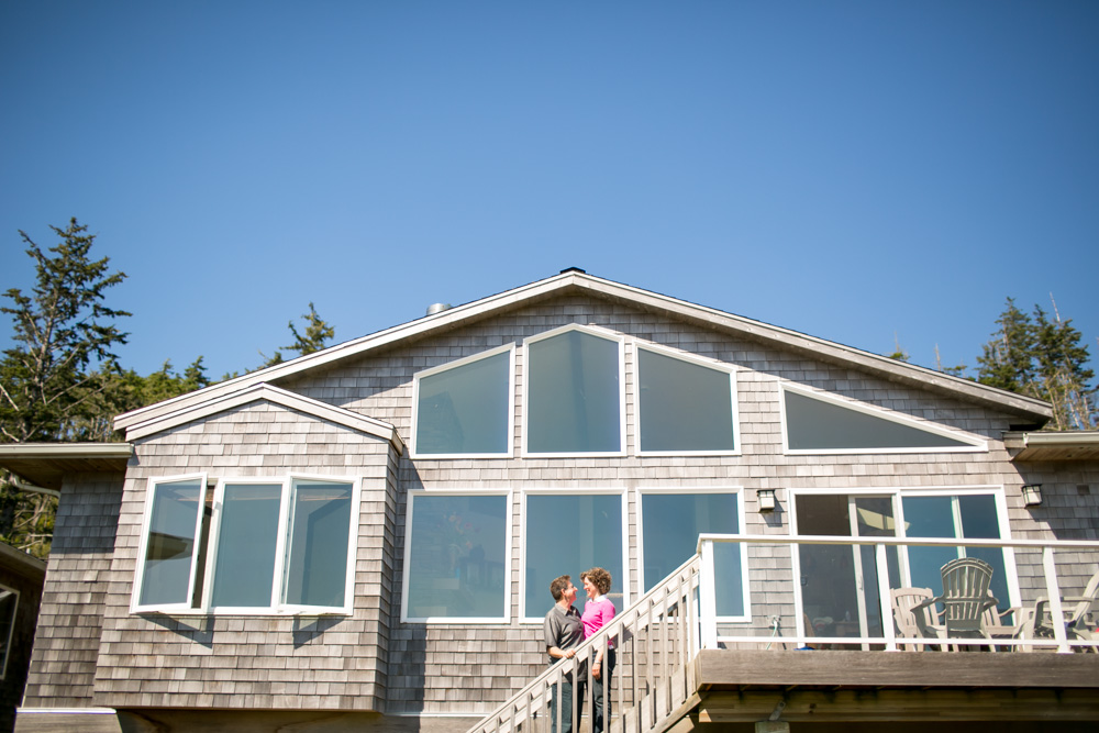 Two people stand together on the outdoor stairs of a wooden house with large windows on a clear, sunny day. Jessica Hill Photography - Wedding Photographer - Oregon