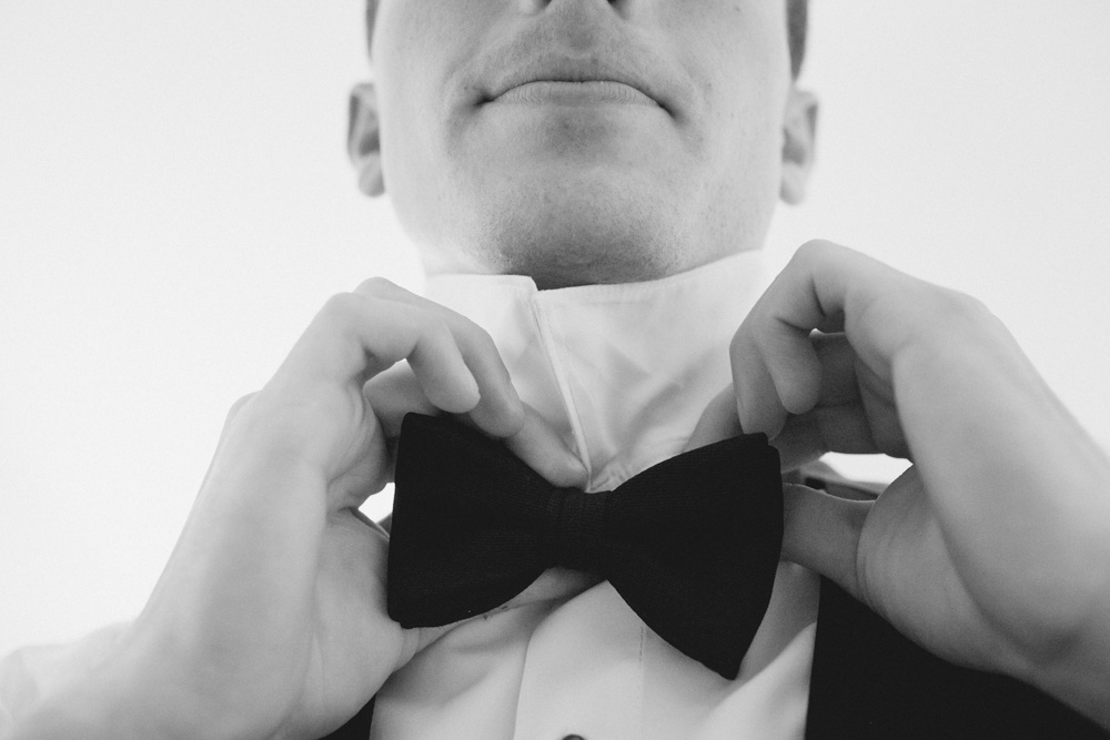 A person adjusts a bow tie on the collar of a dress shirt, seen from the chest up in a black and white photo. Jessica Hill Photography - Wedding Photographer - Oregon