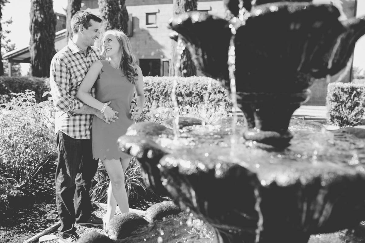A couple stands close together near a fountain in a garden, with the man holding the woman from behind. The image is in black and white. Jessica Hill Photography - Wedding Photographer - Oregon