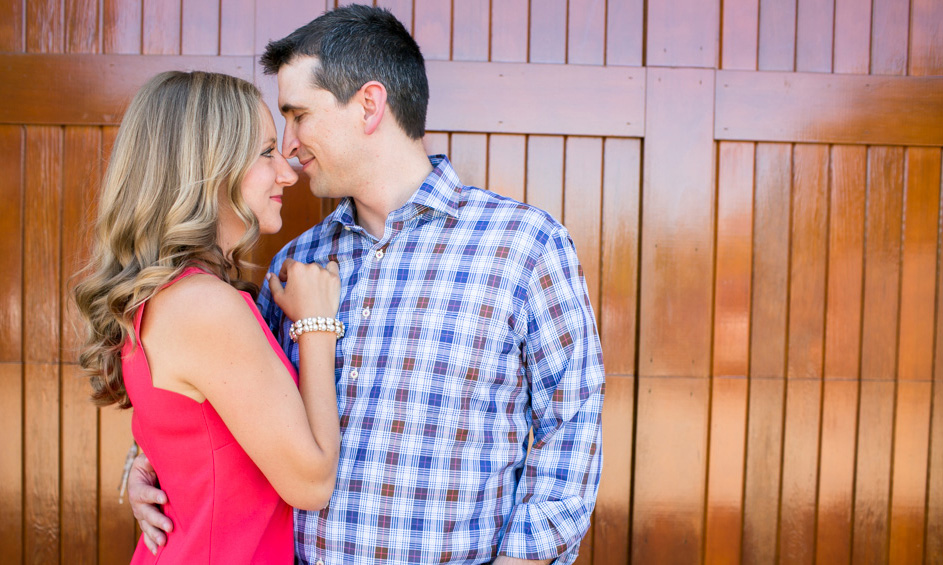 A woman in a pink dress and a man in a plaid shirt stand close together, facing each other and smiling, with a wooden background behind them. Jessica Hill Photography - Wedding Photographer - Oregon