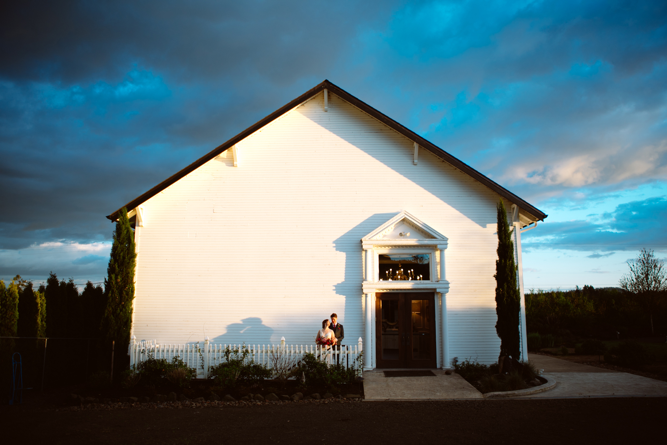 A couple stands outside the entrance of a white building with a pitched roof, illuminated by sunlight, under a partly cloudy sky. Jessica Hill Photography