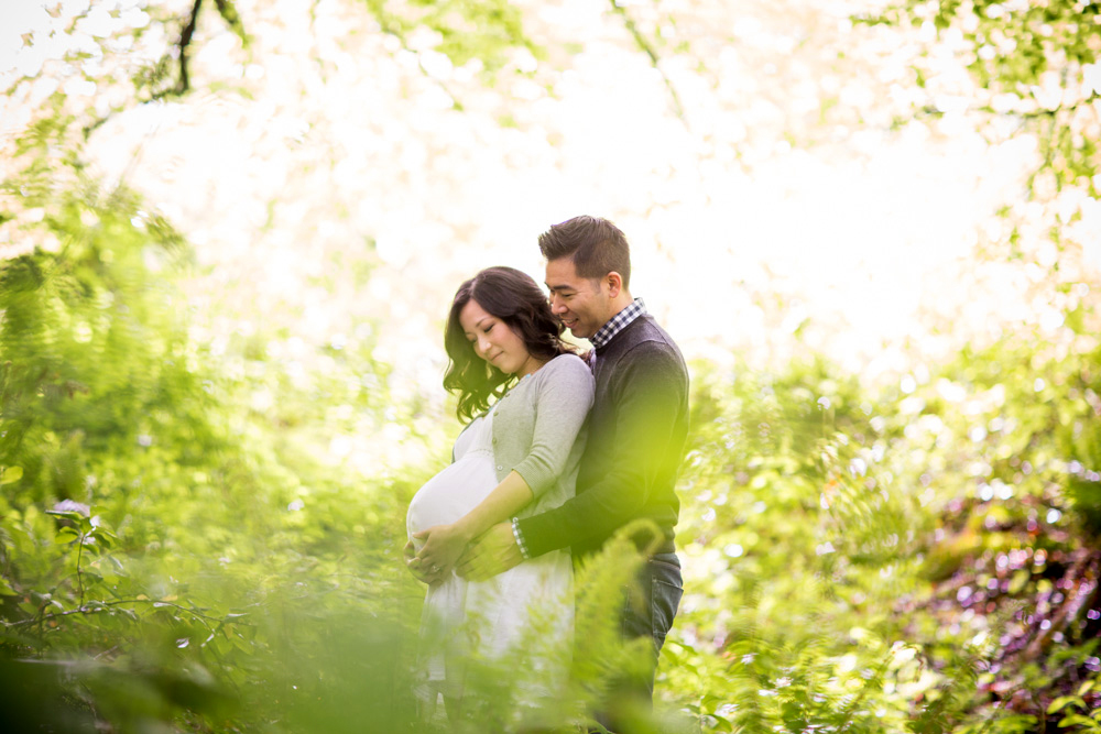 A couple stands outdoors in greenery; the man embraces the pregnant woman from behind as they both look down at her baby bump. Jessica Hill Photography - Wedding Photographer - Oregon
