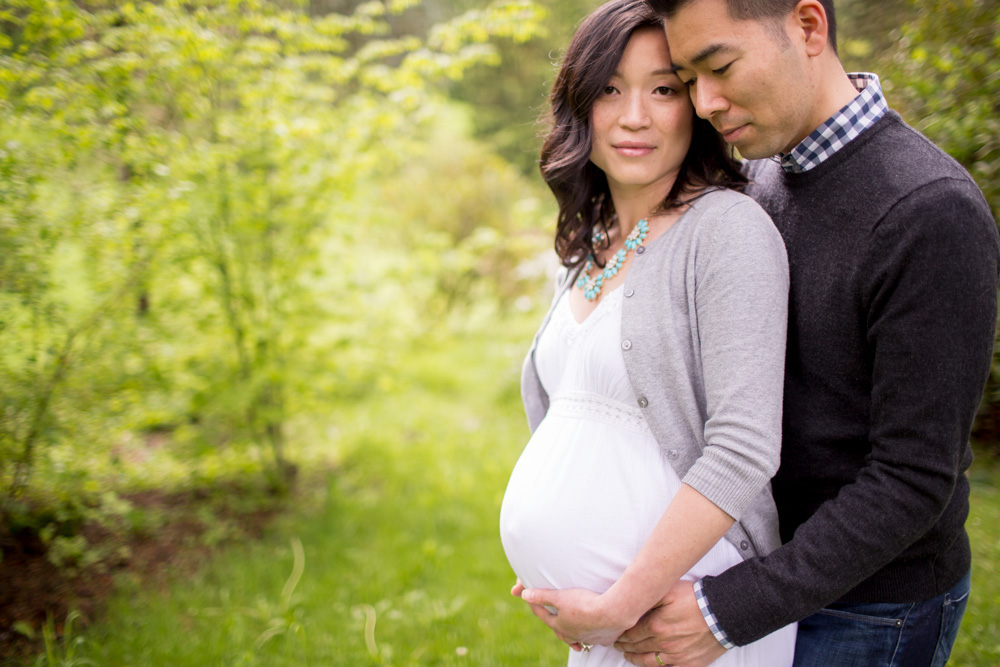 A man stands behind and embraces a pregnant woman outdoors in a green, wooded area. Both are looking at the camera with calm expressions. Jessica Hill Photography - Wedding Photographer - Oregon
