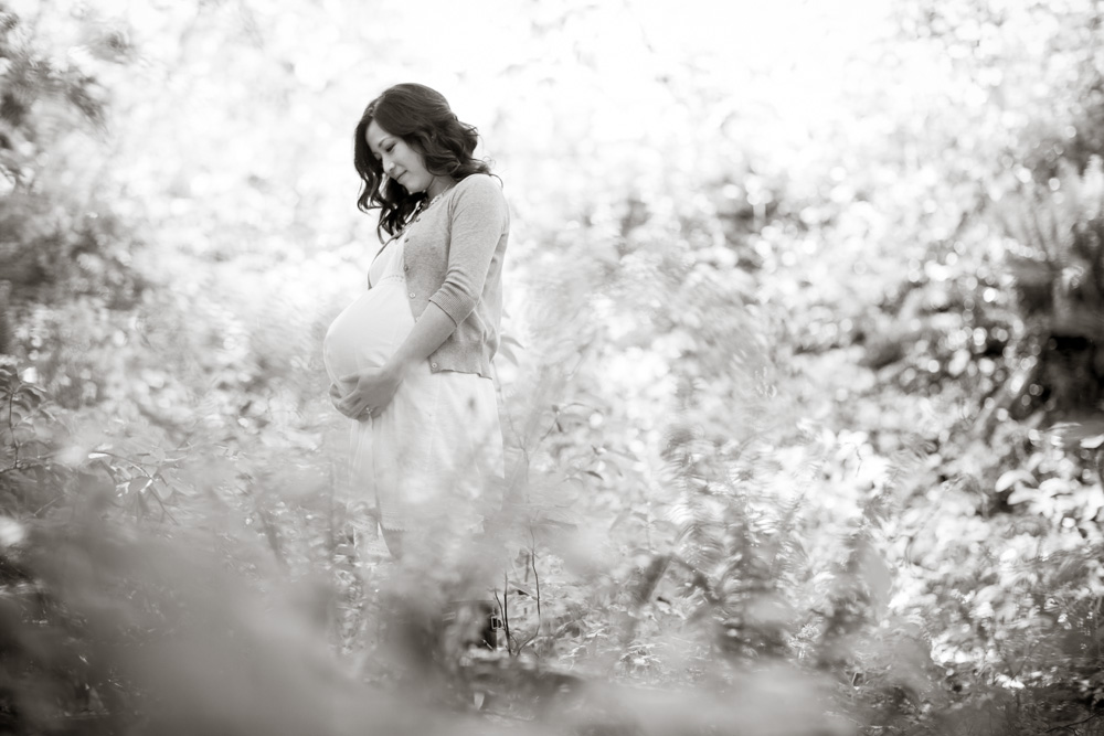 A pregnant woman stands outdoors in a light dress and sweater, looking down and holding her belly, surrounded by blurred foliage. Jessica Hill Photography - Wedding Photographer - Oregon