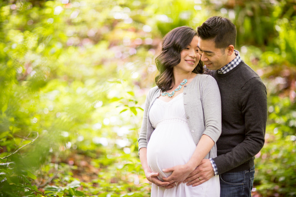 A pregnant woman in a white dress stands outdoors with a man behind her, both smiling, in a green, leafy setting. Jessica Hill Photography - Wedding Photographer - Oregon
