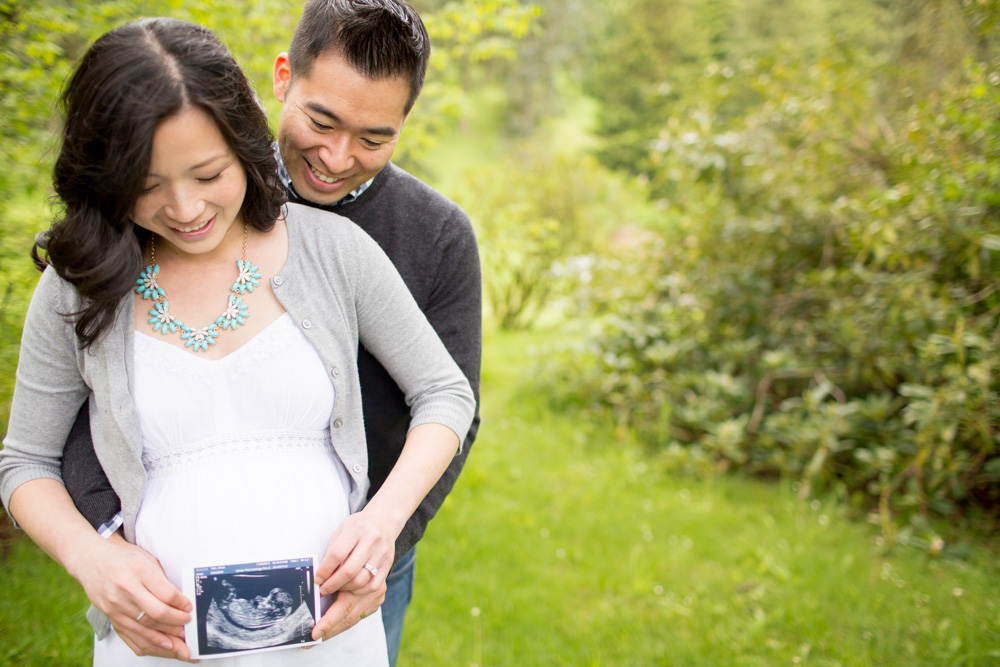 A couple stands outside, smiling, as the woman holds an ultrasound photo in front of her pregnant belly. Jessica Hill Photography - Wedding Photographer - Oregon