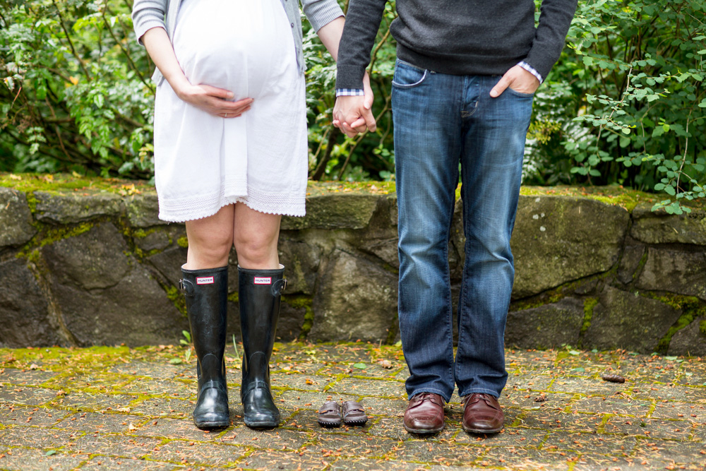 A pregnant woman in a white dress and rain boots stands holding hands with a man in jeans; baby shoes are placed on the ground between them. Jessica Hill Photography - Wedding Photographer - Oregon