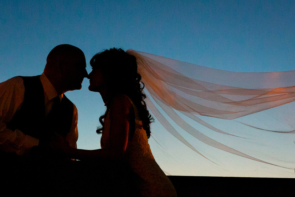 Silhouetted bride and groom kiss at dusk, with the bride’s veil flowing behind her against a clear blue sky. Jessica Hill Photography - Wedding Photographer - Oregon