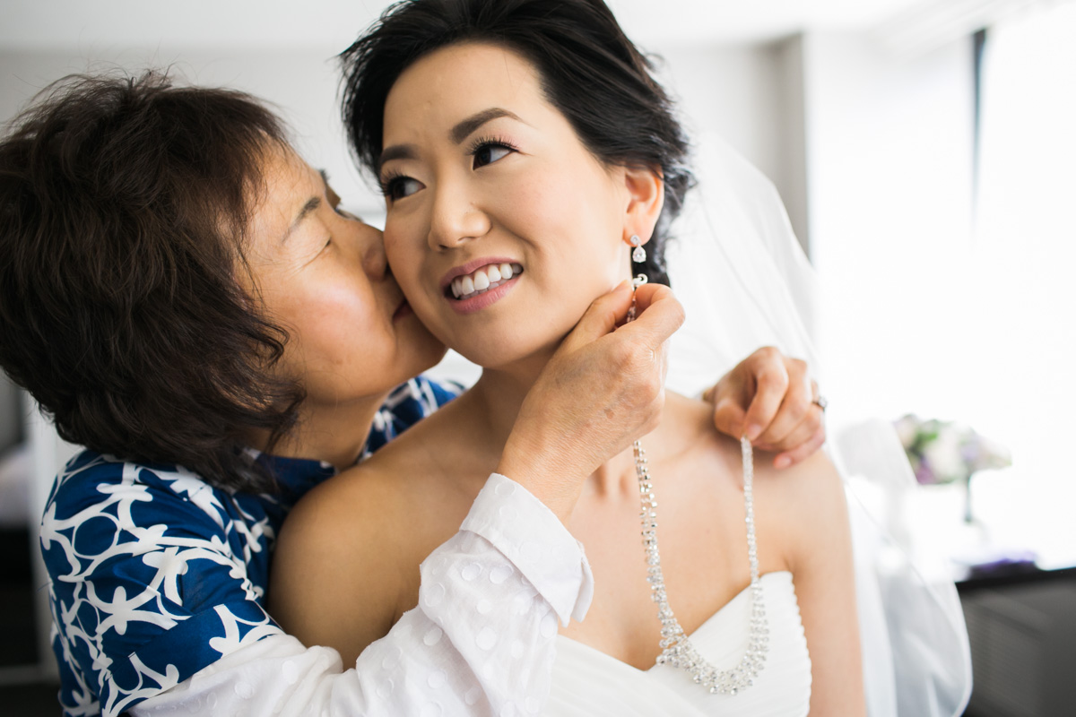 A woman in a blue and white blouse kisses a smiling bride on the cheek while helping her put on a necklace. The bride, wearing a strapless white dress and veil, looks happy and touched. Jessica Hill Photography - Wedding Photographer - Oregon