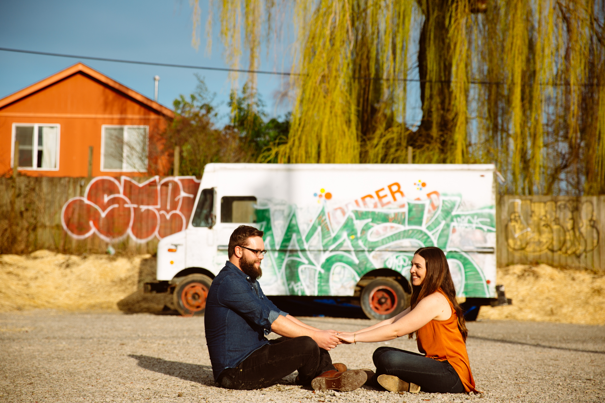 A woman in a light blue button-up shirt smiles and looks down in a bright room, with two people and various household items blurred in the background. Jessica Hill Photography - Wedding Photographer - Oregon