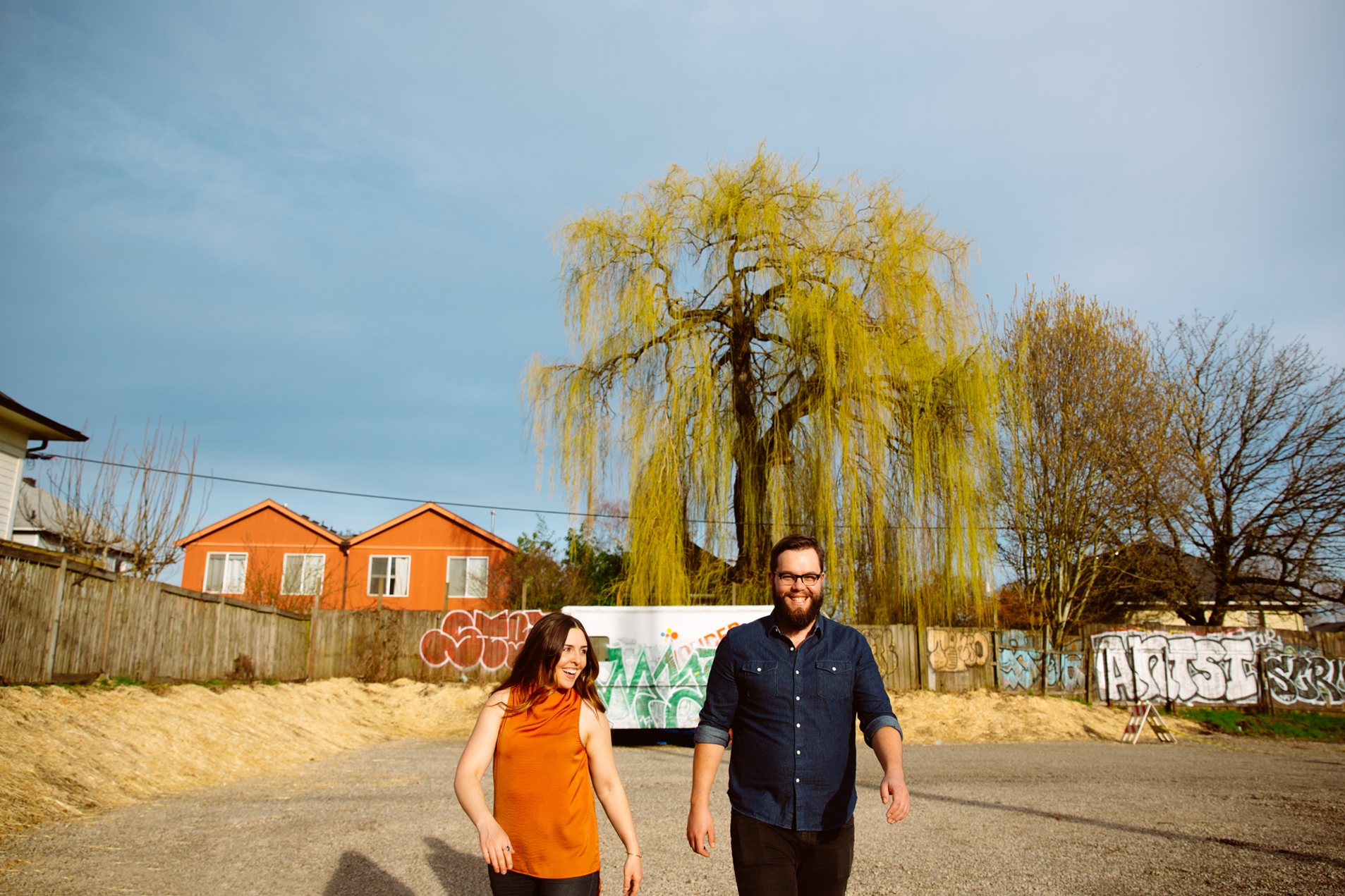 A man adjusts his black suspenders over a white dress shirt, wearing a gold watch and wedding ring. In the background, another man in formalwear is crouched, tying his shoes in a living room with a sofa and patterned pillows. Jessica Hill Photography - Wedding Photographer - Oregon