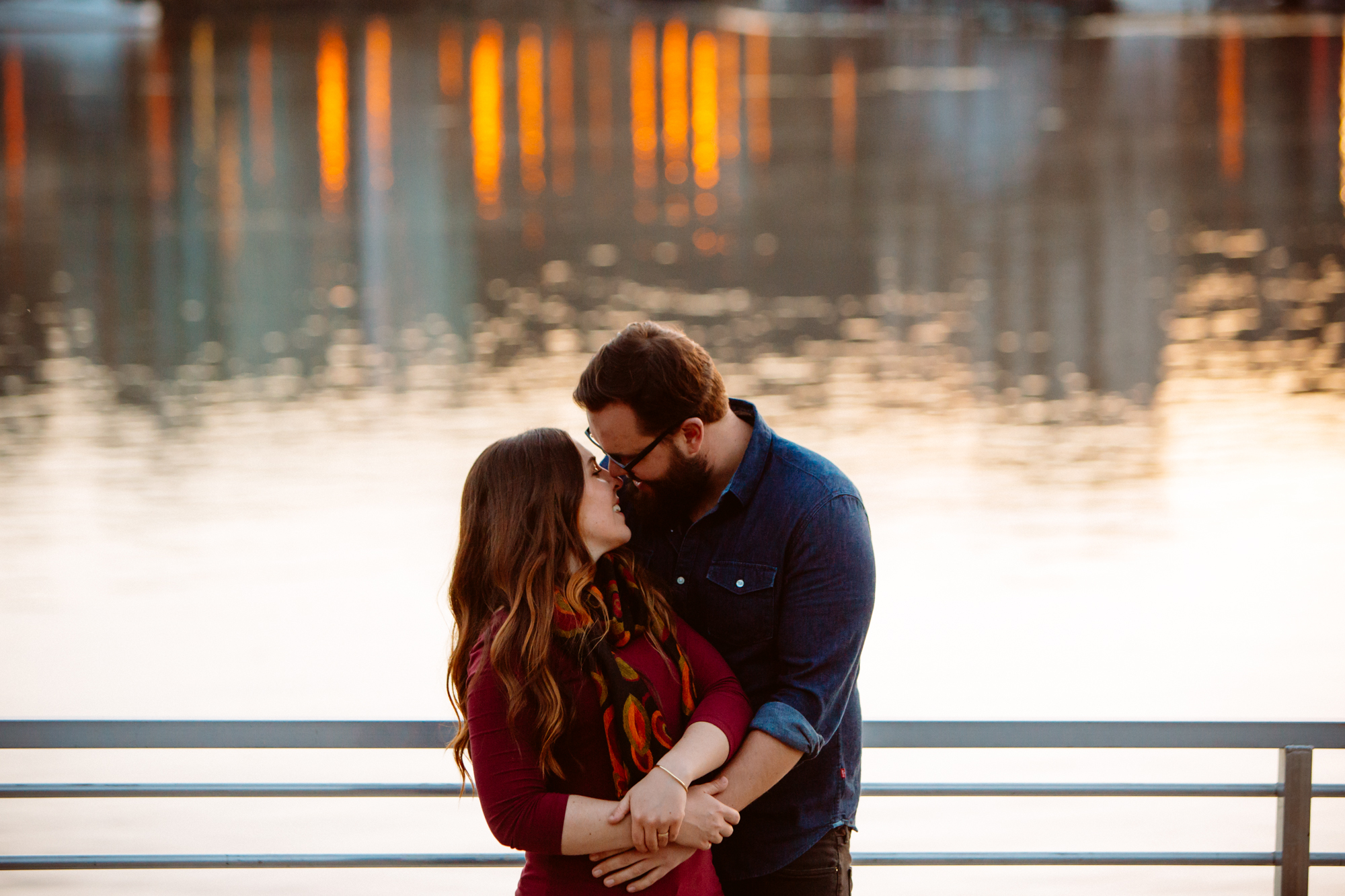 A bride and groom walk hand in hand along a rocky shoreline at sunset, with calm water and tree-covered mountains in the background. Jessica Hill Photography - Wedding Photographer - Oregon