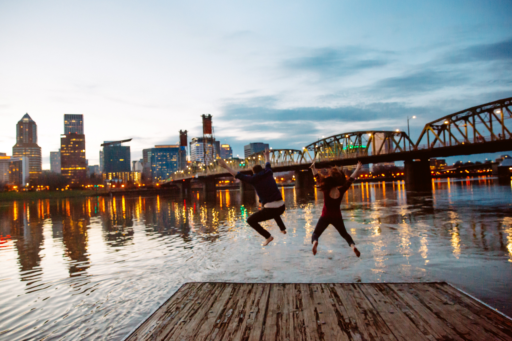 A black-and-white photo of a couple embracing closely in front of tall grass, with modern high-rise buildings blurred in the background. The scene feels romantic and intimate. Jessica Hill Photography - Wedding Photographer - Oregon