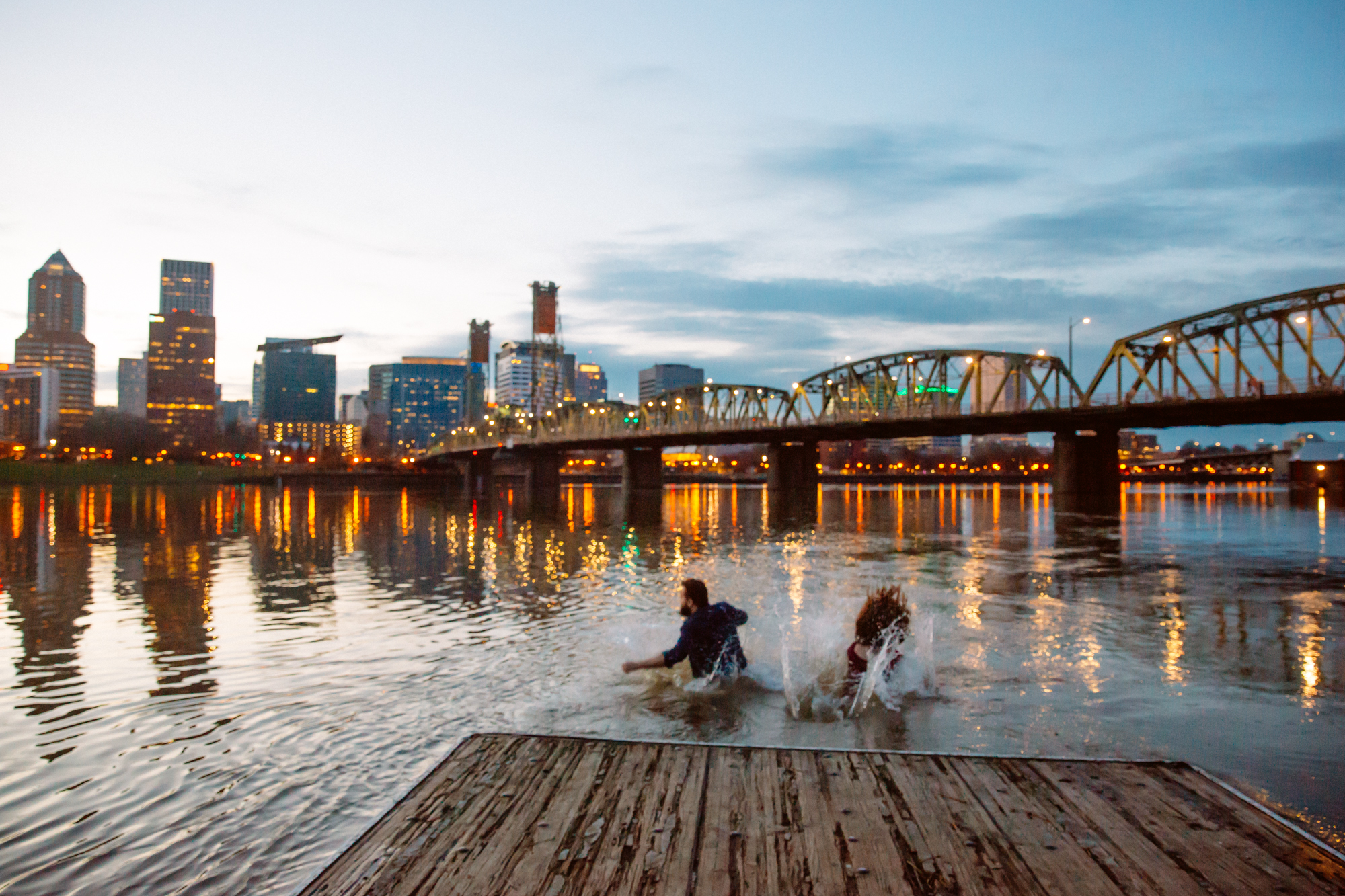 A bride and groom walk arm in arm down a sunlit path in a grassy field, surrounded by trees and mountains, smiling and dressed in formal wedding attire. Jessica Hill Photography - Wedding Photographer - Oregon