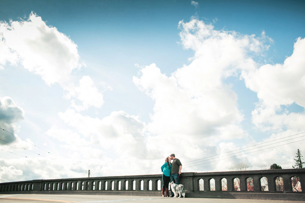 A couple stands close together on a bridge with a dog at their feet, under a sky filled with scattered clouds. Jessica Hill Photography - Wedding Photographer - Oregon