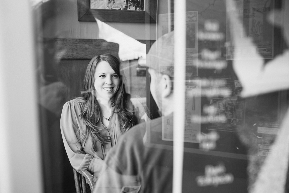 A woman sitting at a table smiles and talks to a man across from her, seen through a glass window in a black-and-white photo. Jessica Hill Photography - Wedding Photographer - Oregon