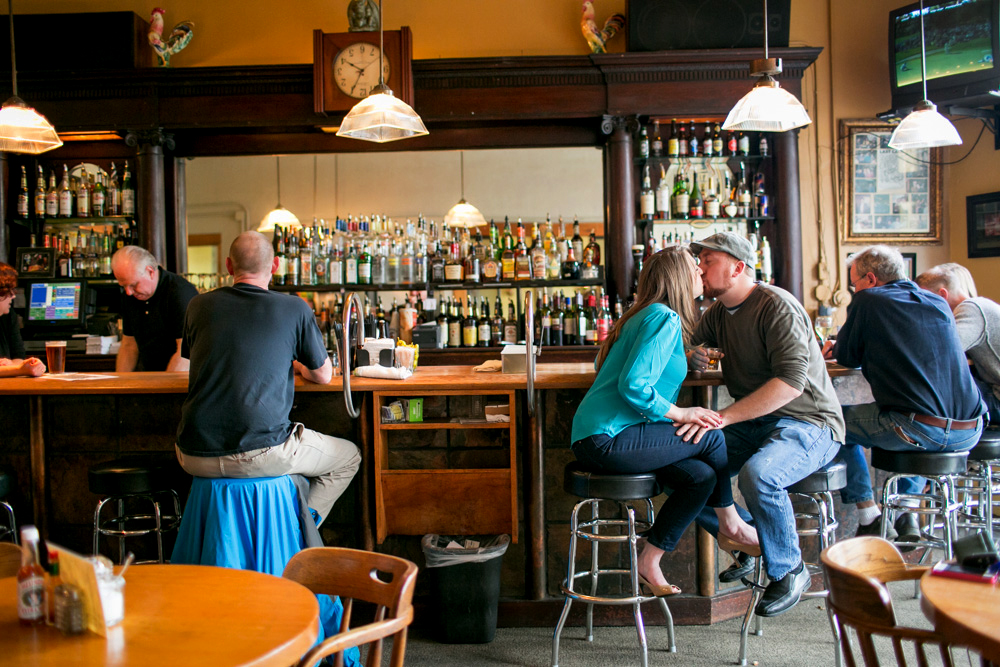 People sit at a bar; a couple on the right shares a kiss while others converse or face the bartender. Bottles, glasses, and lights are visible behind the bar. Jessica Hill Photography - Wedding Photographer - Oregon